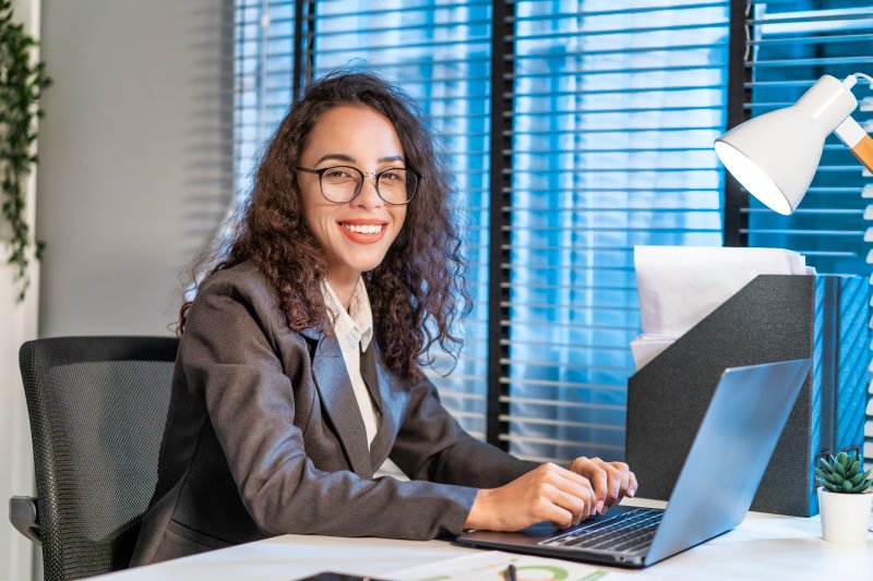 Mujer joven vestida para la oficina sentada frente a un computador, mostrándose feliz con su trabajo.